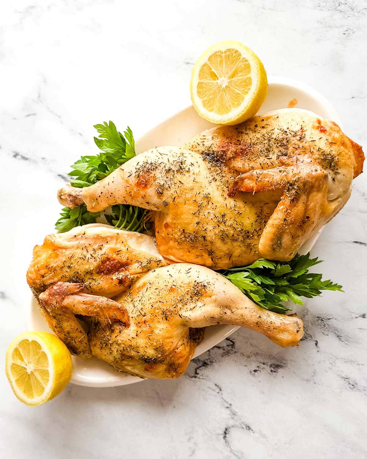 Two halves of a roasted chicken on a white platter with parsley and lemons.