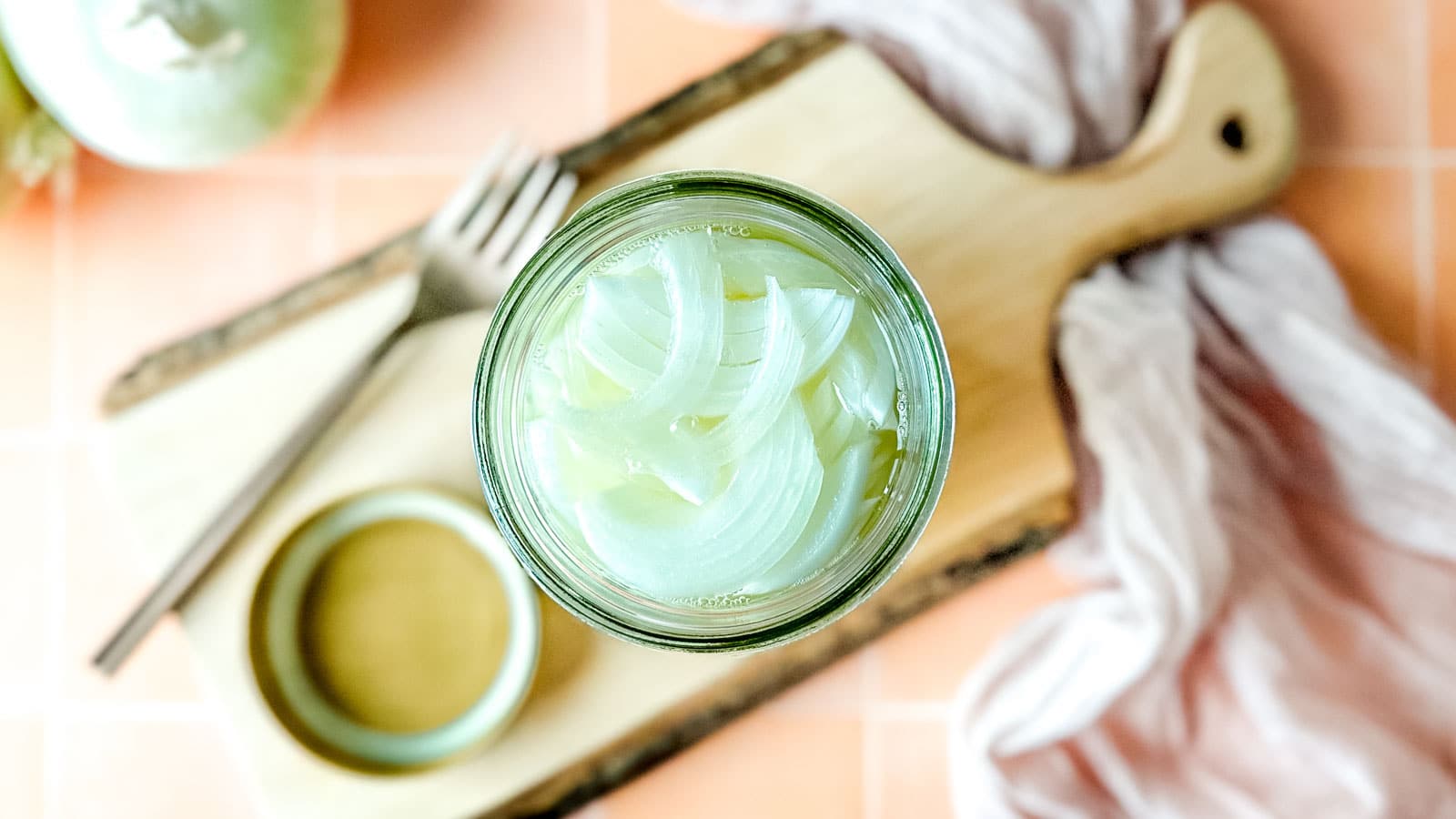 Pickled white onions in a glass jar on a wooden cutting board surrounded by white onions, a fork, and a pink linen.