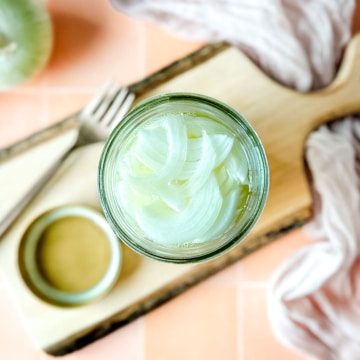 Pickled white onions in a glass jar on a wooden cutting board surrounded by white onions, a fork, and a pink linen.