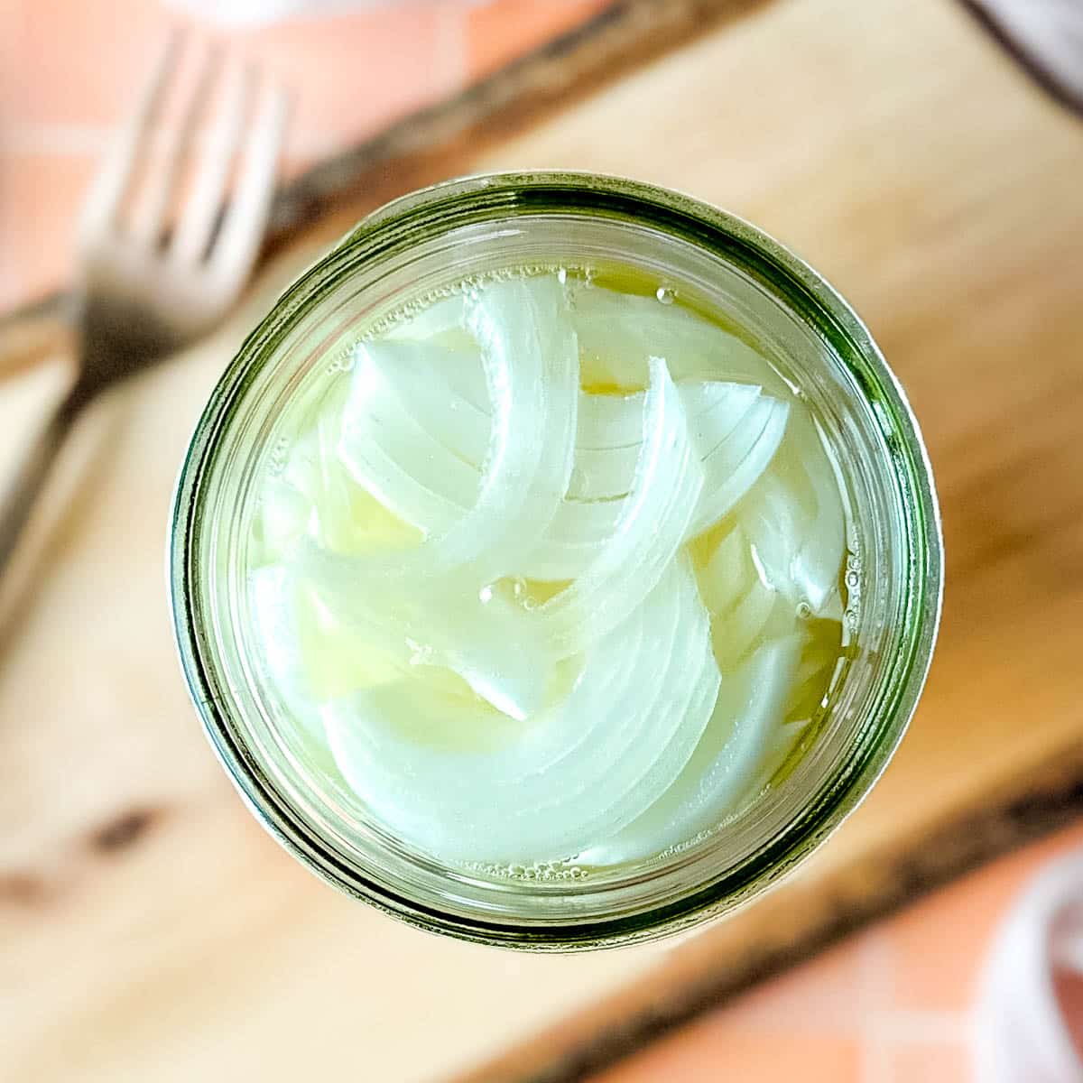 Pickled white onions in a glass jar on a wooden cutting board surrounded by white onions, a fork, and a pink linen.