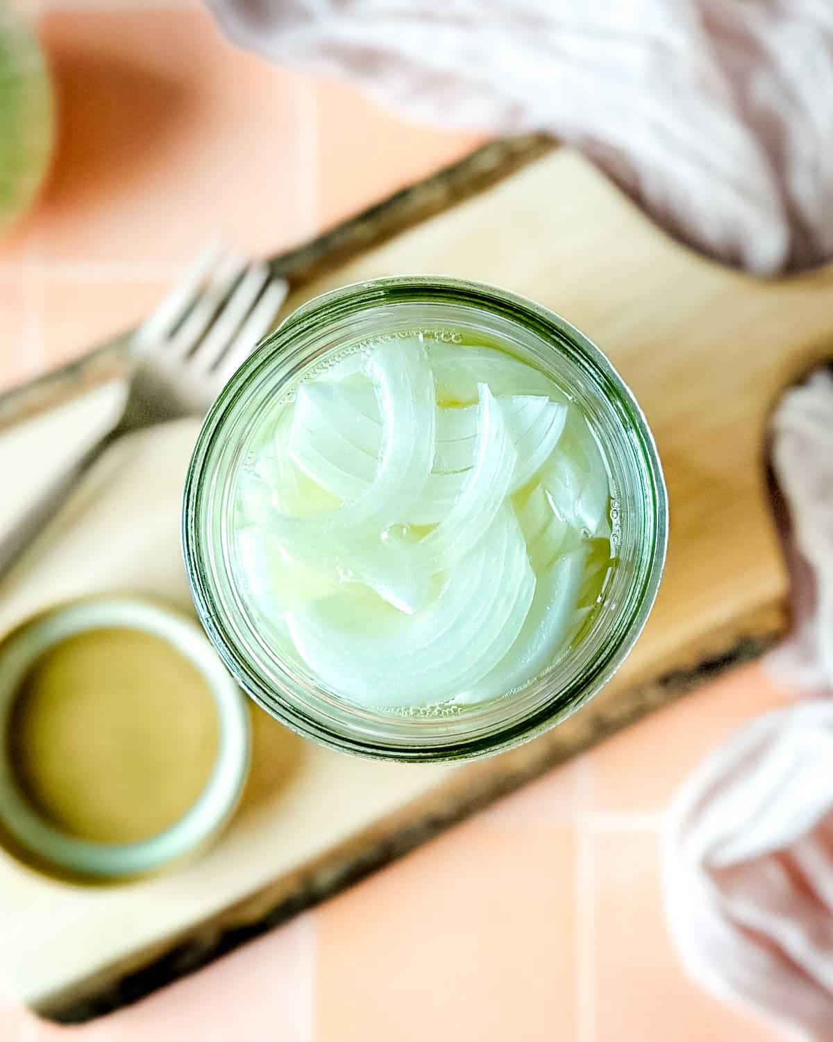 Pickled white onions in a glass jar on a wooden cutting board surrounded by white onions, a fork, and a pink linen.