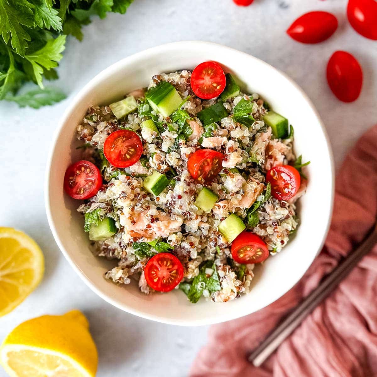 Salmon quinoa salad in a white bowl surrounded by cherry tomatoes, fresh parsley, and a cut lemon.