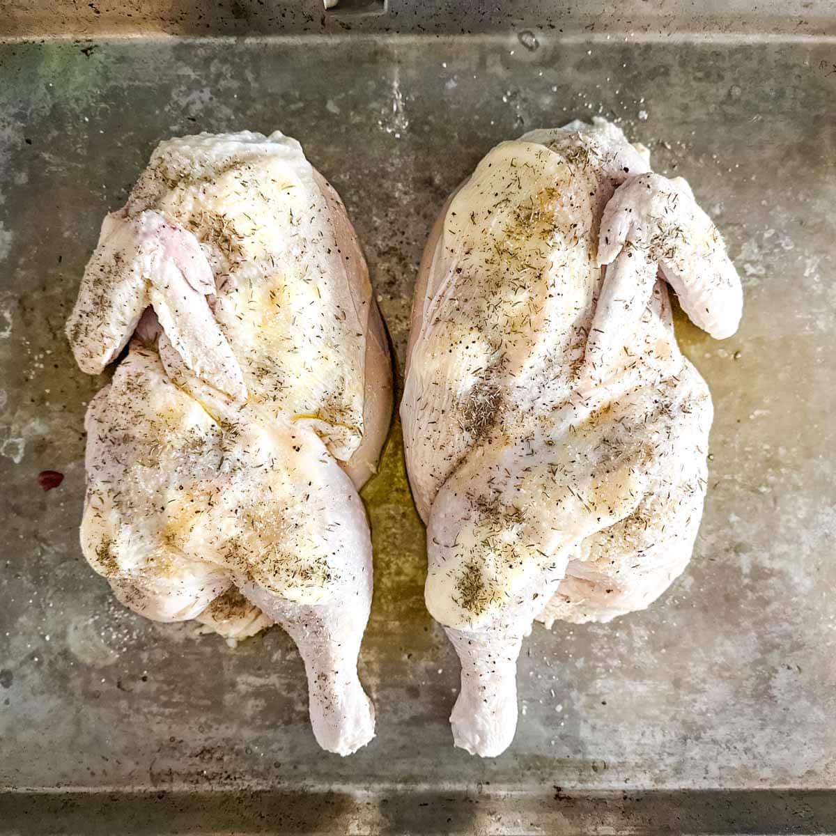 Two halves of a chicken on a sheet pan seasoned with olive oil, salt and pepper, thyme, and garlic.