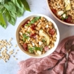Whole wheat pasta salad in a white bowl surrounded by fresh basil, pine nuts, and a pink linen.