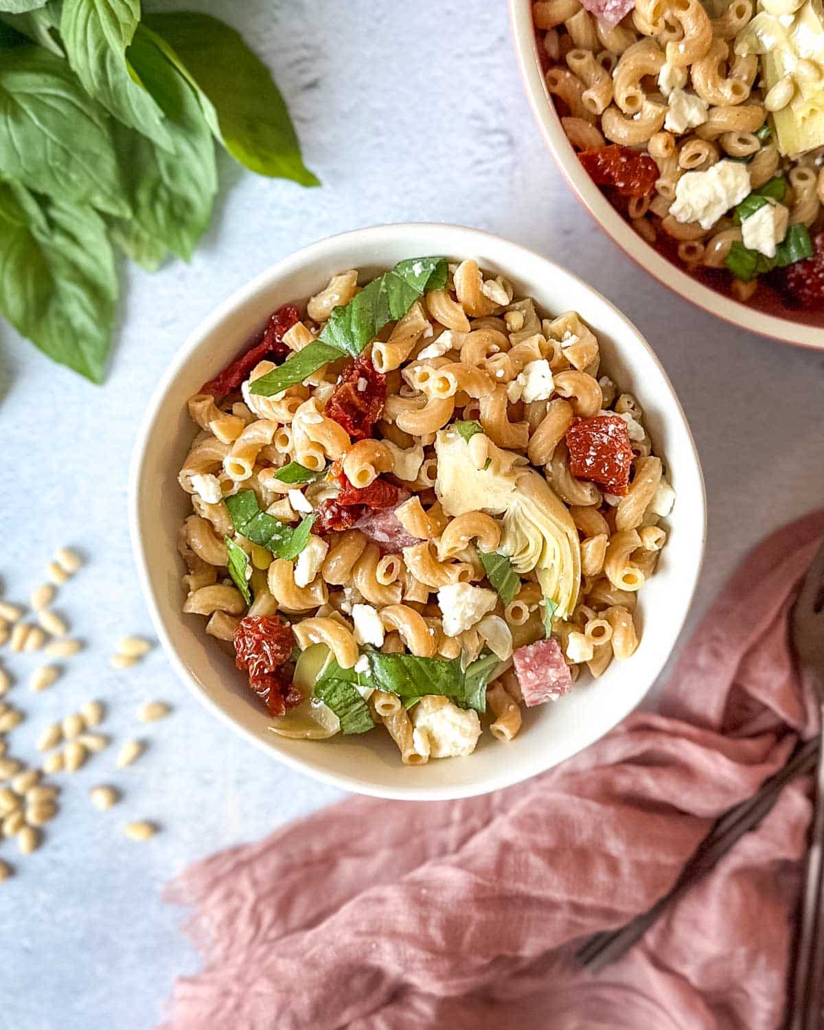 Whole wheat pasta salad in a white bowl surrounded by fresh basil, pine nuts, and a pink linen.