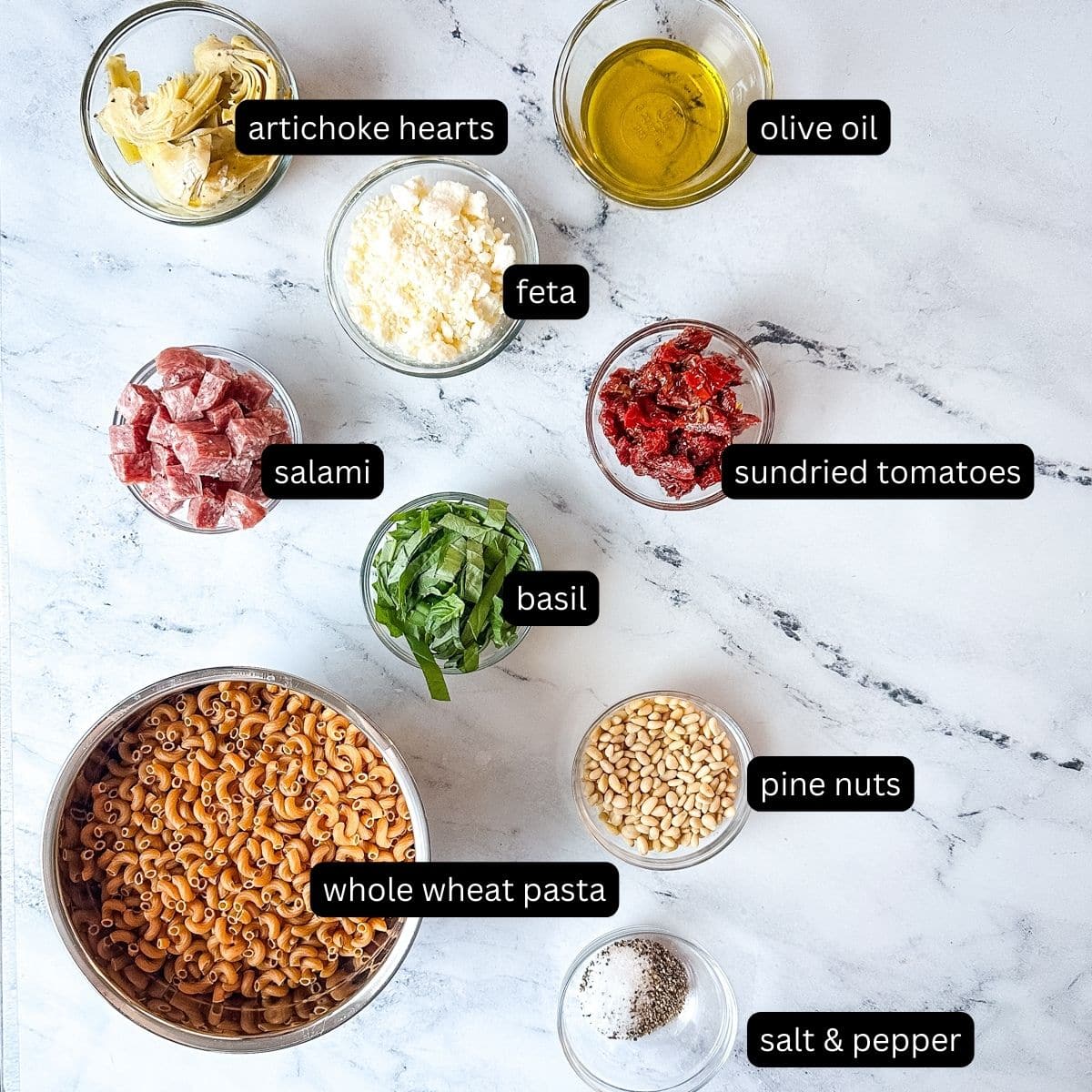 Labeled ingredients for whole wheat pasta salad on a white marble counter.