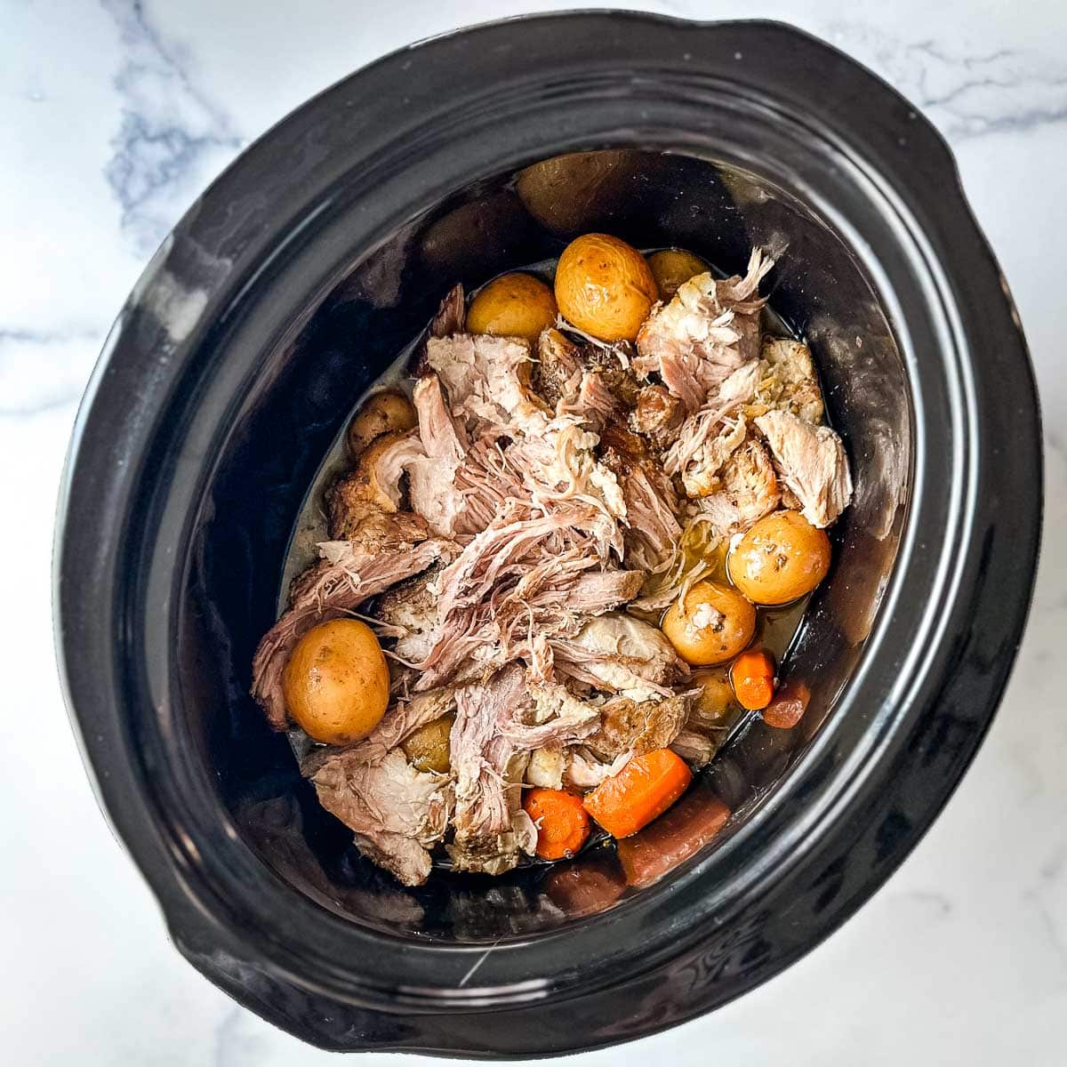 A crock pot full of shredded pork roast, potatoes and carrots.