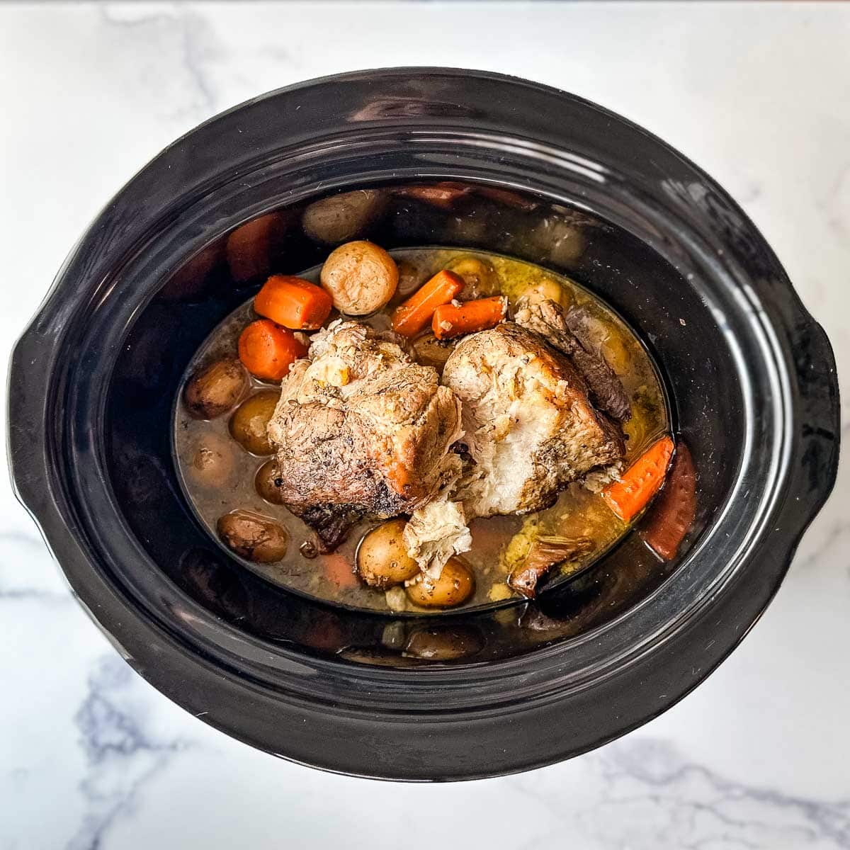 A crock pot containing a fully cooked Italian slow cooker pork roast, potatoes, and carrots.
