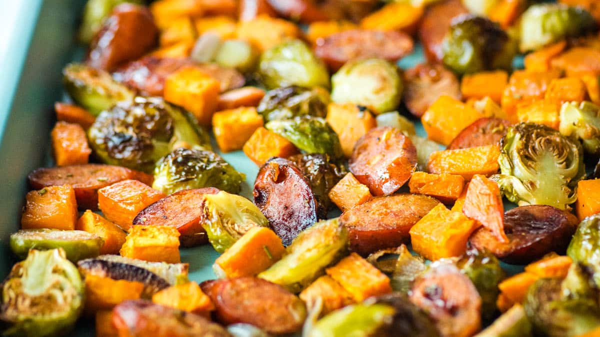 Roasted brussels sprouts, sweet potatoes, shallots, and chicken sausage on a baking sheet.
