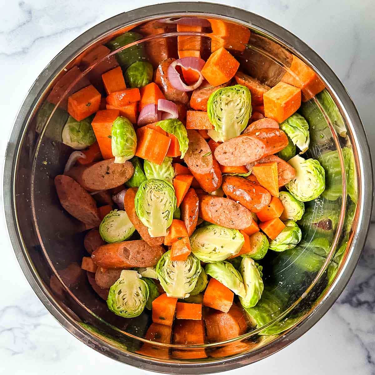 Brussels sprouts, sweet potato, shallots, rosemary, and sausage in a bowl.