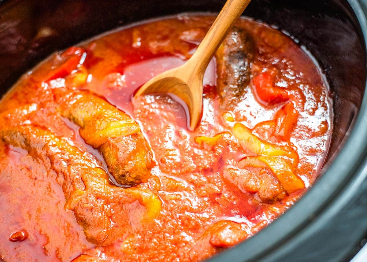 A wooden spoon in a slow cooker with sausage, peppers, and marinara sauce.