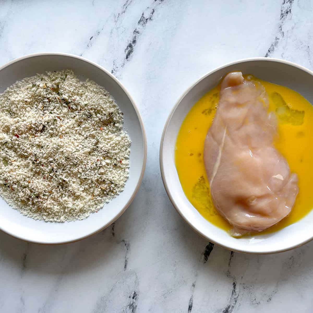 One bowl with panko breadcrumbs and one bowl with beaten egg and a chicken breast on a marble counter.