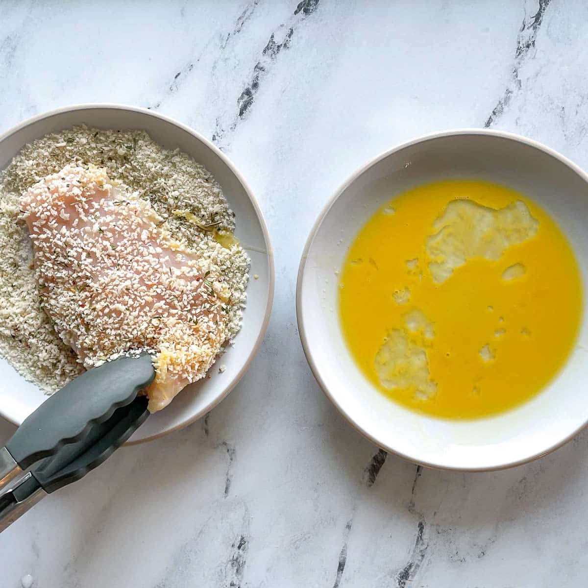 One bowl with panko breadcrumbs and a breaded chicken cutlet and one bowl with beaten egg on a marble counter.