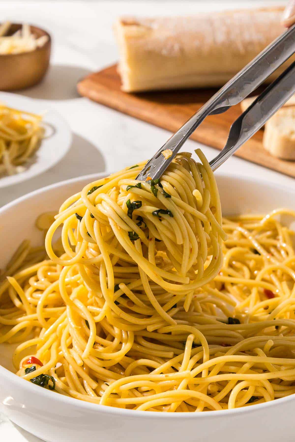 A person is holding a pair of tongs over a bowl of spaghetti aglio olio e peperoncino.