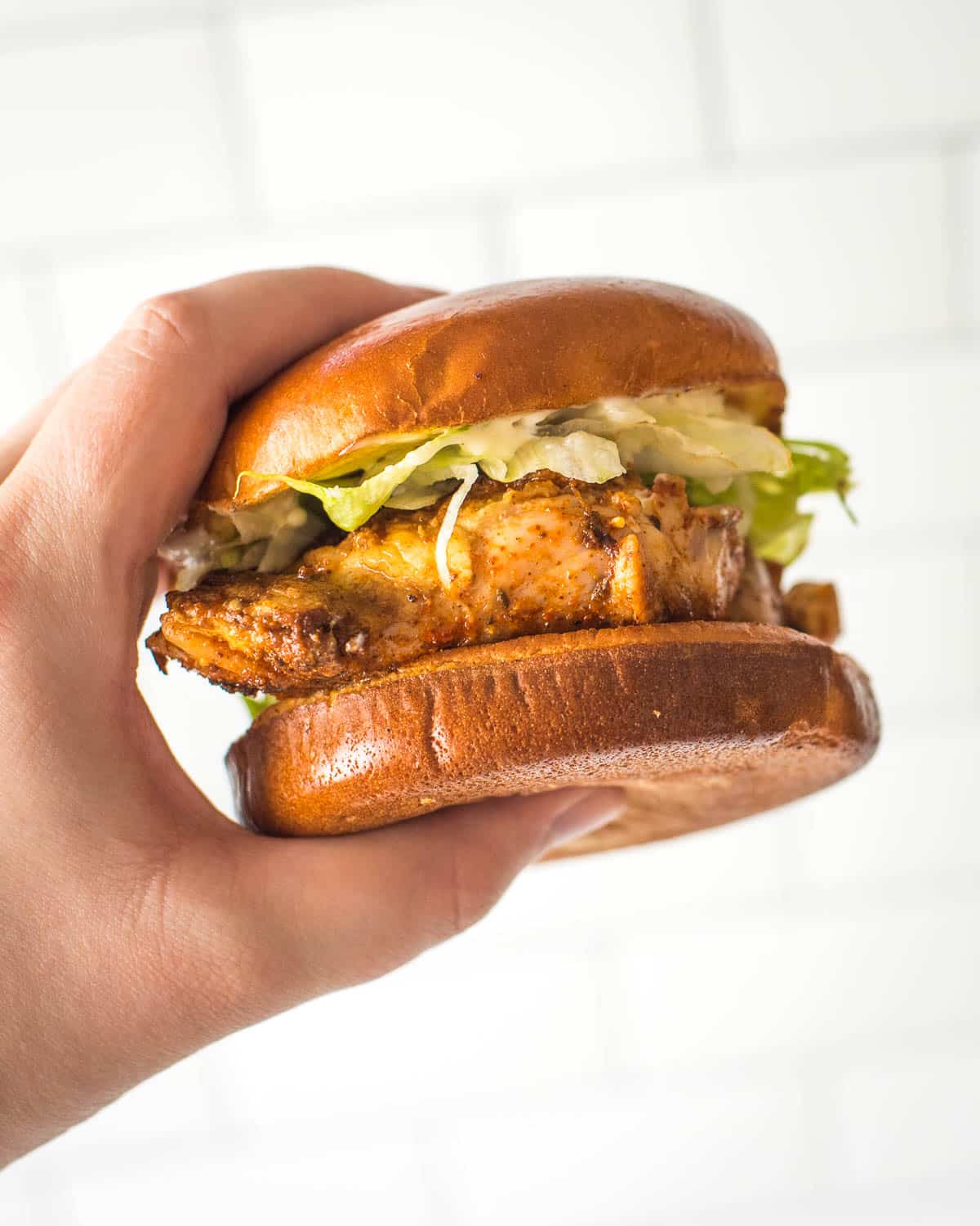 A person holding a blackened chicken sandwich in front of a tiled wall.