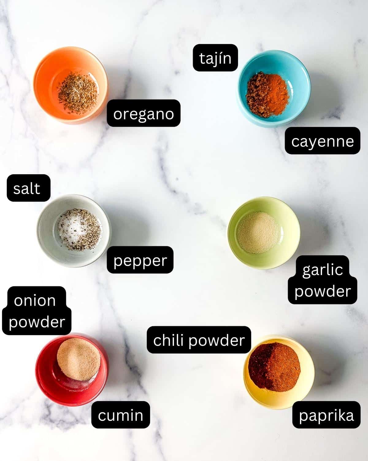 Labeled spices and seasonings for taco seasoning in bowls on a marble countertop.