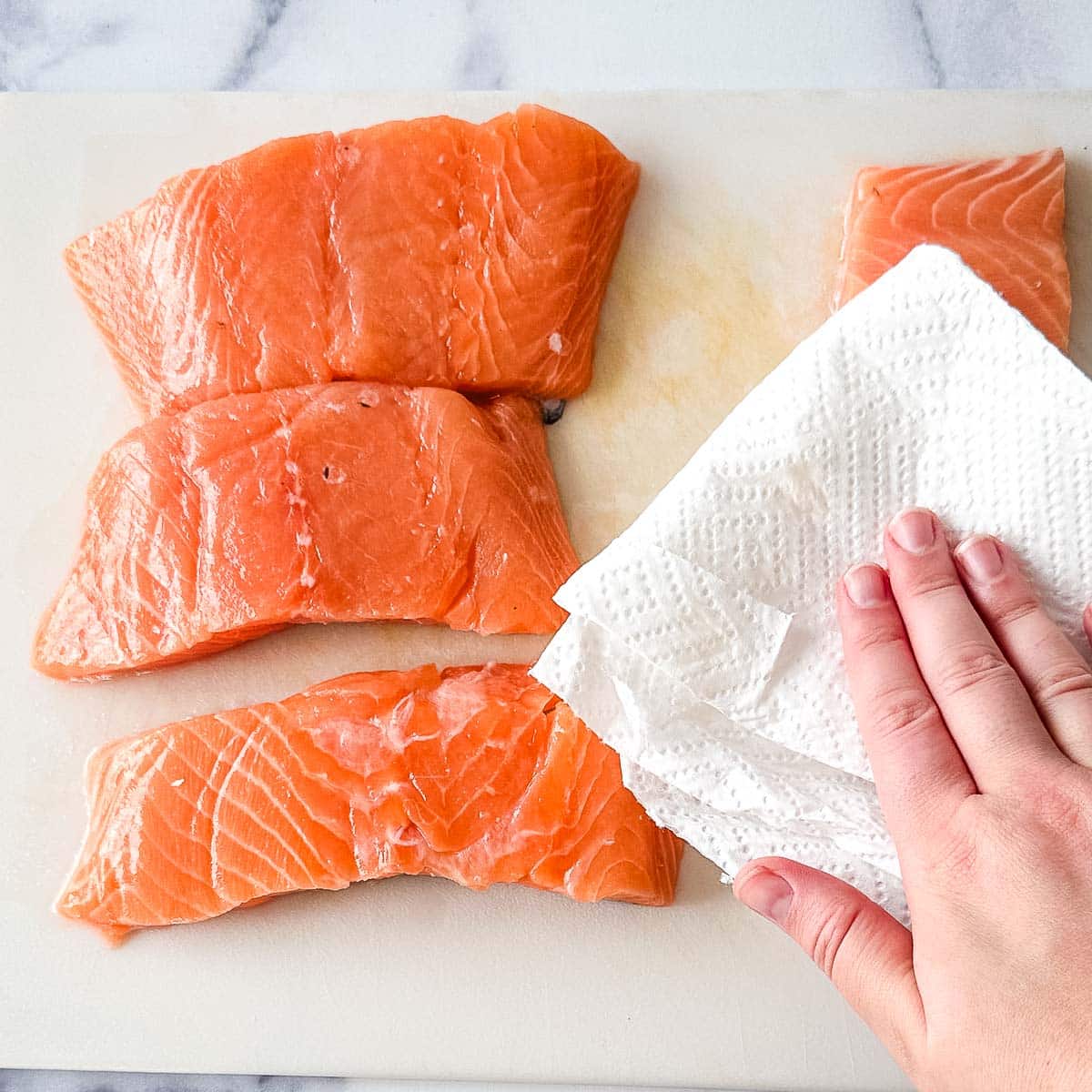 Salmon fillets on a cutting board with a hand patting them dry them with a paper towel.