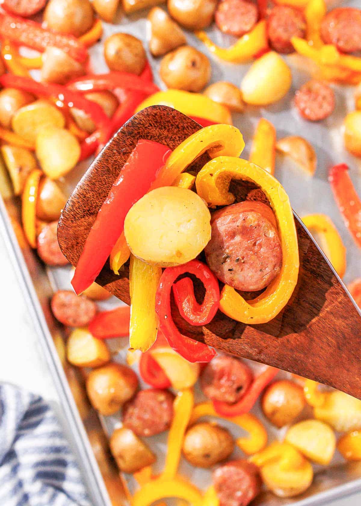 A spoon holds a bite of chicken sausage, peppers, and potatoes above a sheet pan with potatoes, chicken sausage and peppers on it.