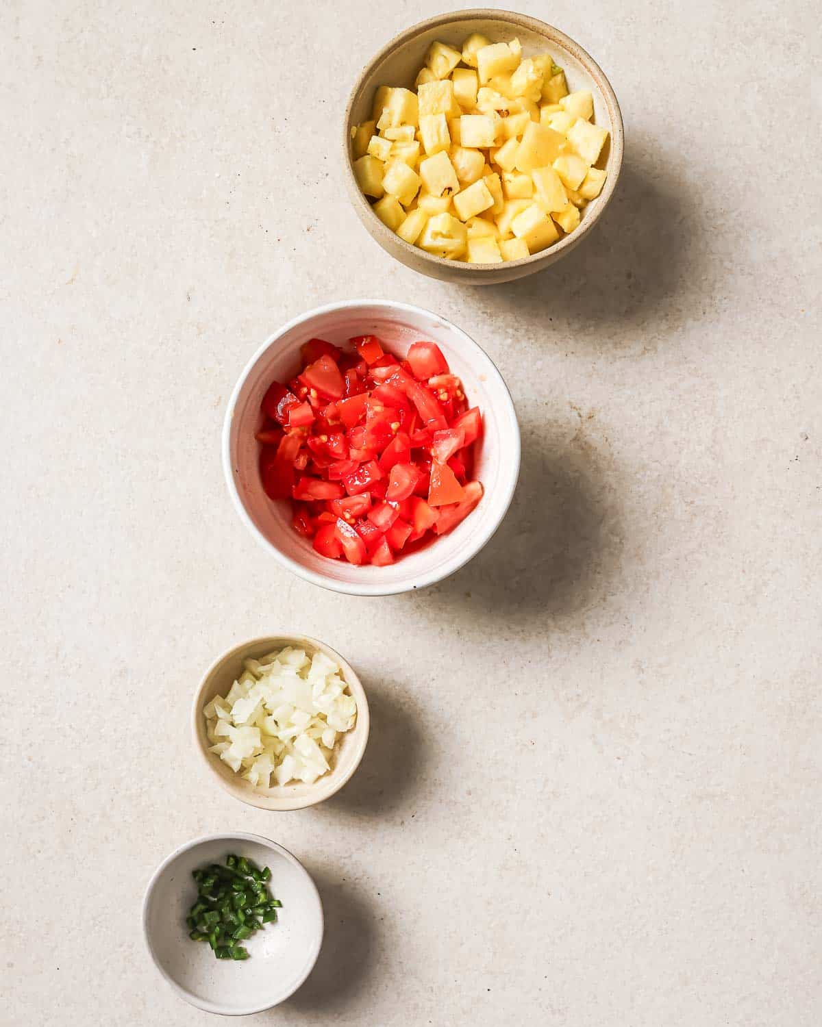 Several bowls containing the chopped ingredients for pineapple salsa on a white surface.