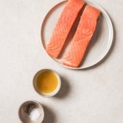 Two pieces of salmon on a plate with salt, pepper, and oil.
