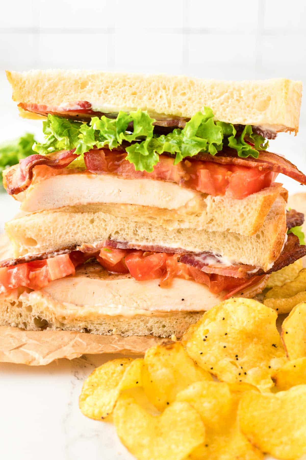 A sandwich with bacon, lettuce, tomatoes, chicken next to potato chips.