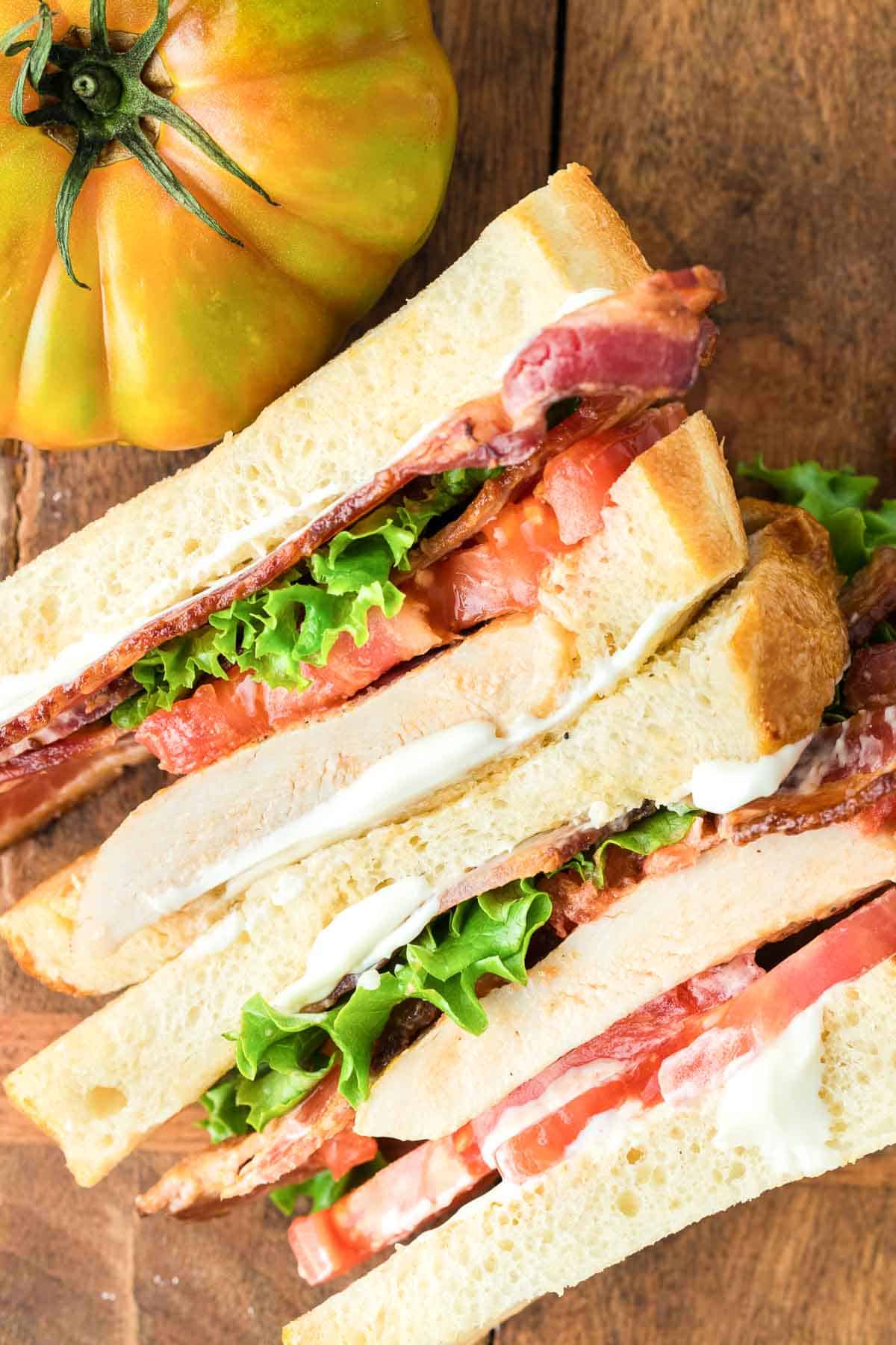 A sandwich with bacon, tomatoes, chicken, and lettuce on a wooden cutting board.