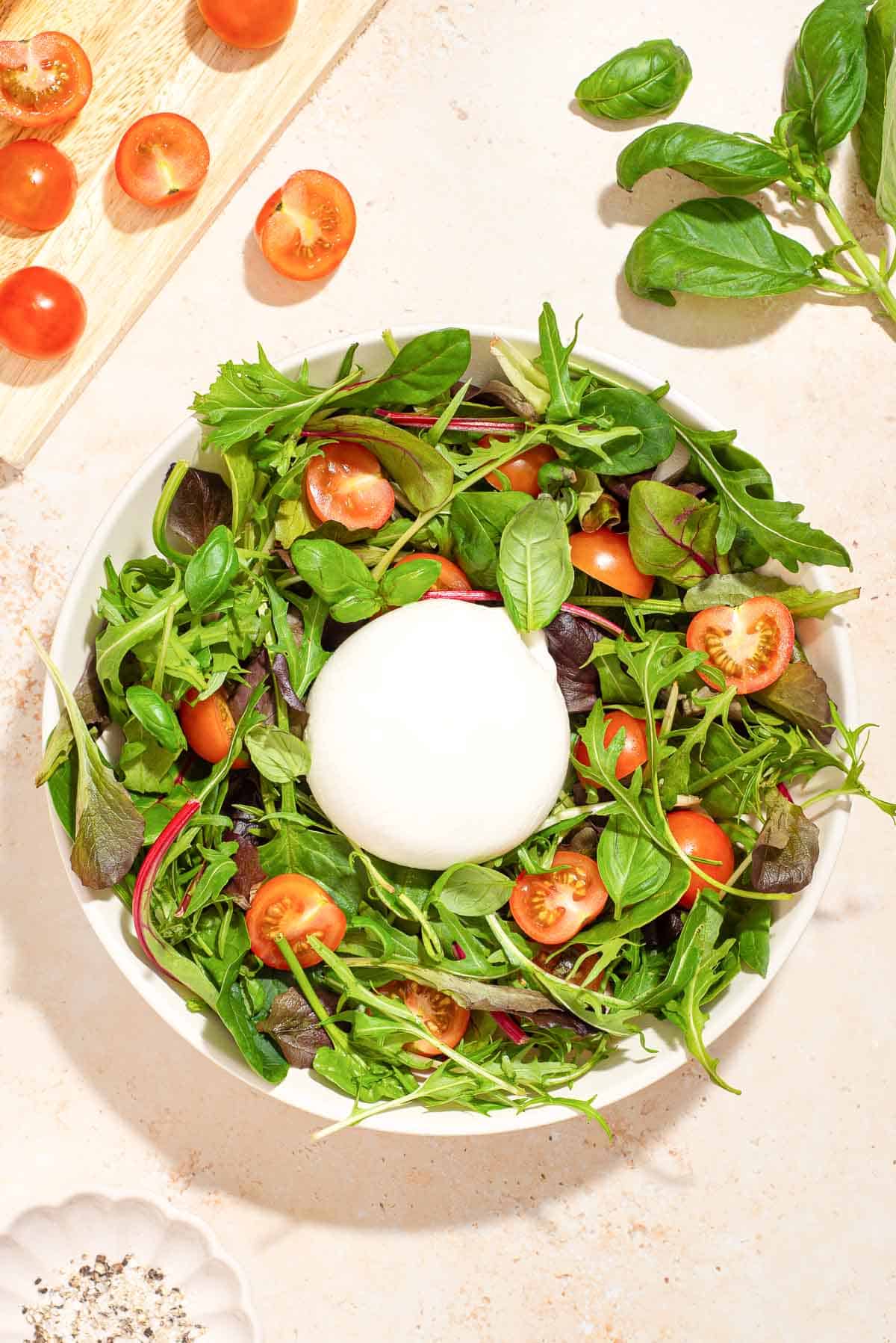 A white bowl with greens, tomatoes, and burrata in it.