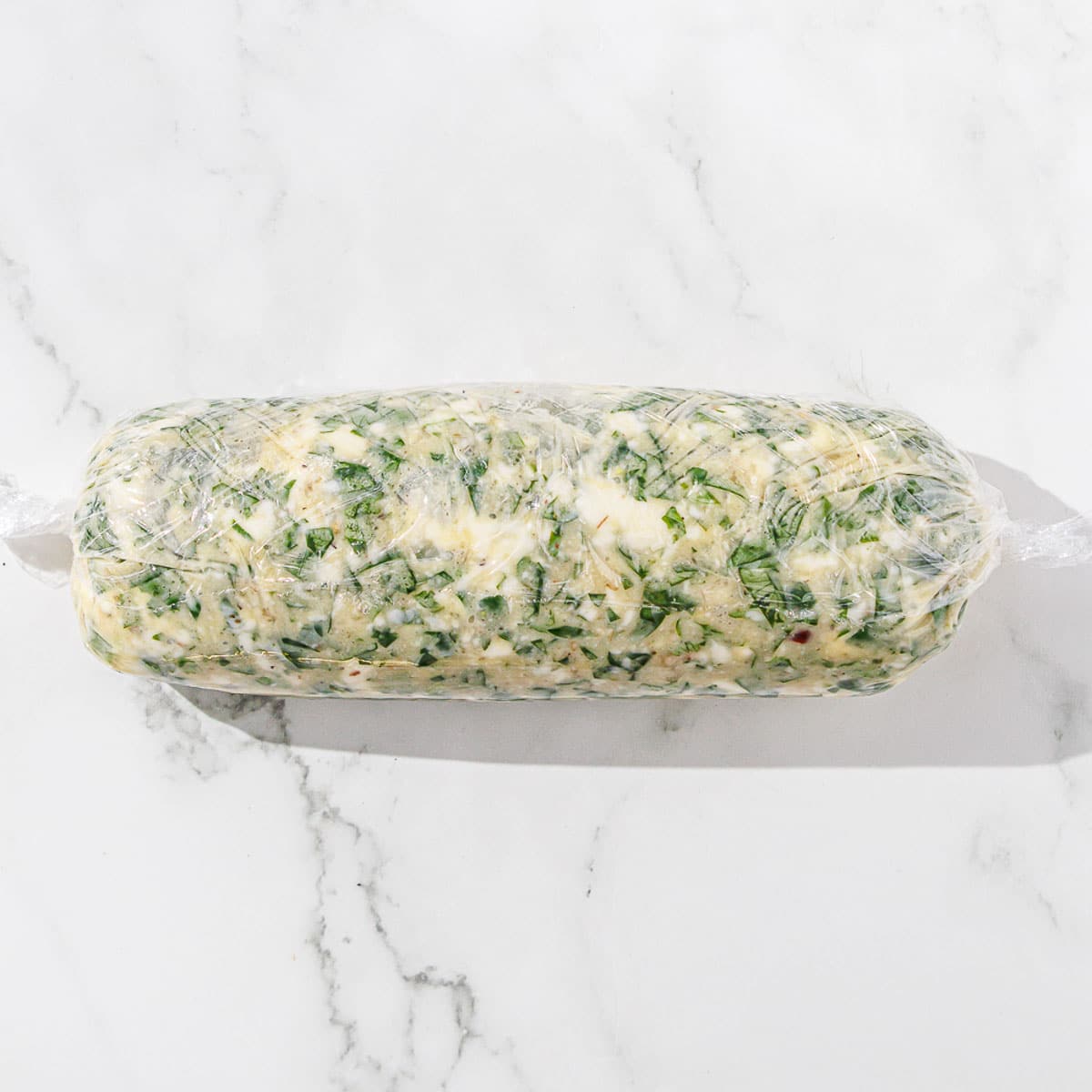 A roll of chimichurri butter wrapped in plastic on a marble surface.