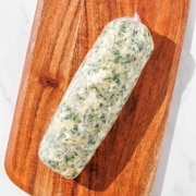 A log of chimichurri butter wrapped in plastic wrap on a wooden cutting board.