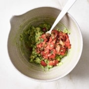 Salsa is added to guacamole in a white bowl with a spoon.