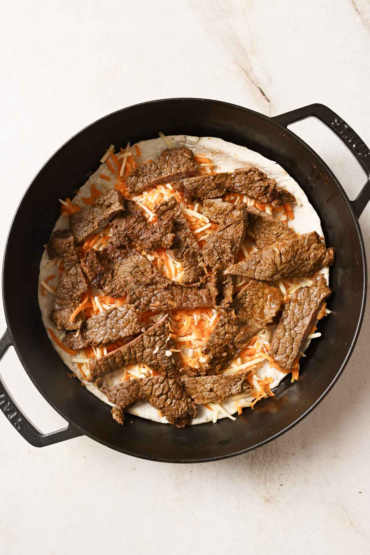 A skillet with a tortilla, cheese, and sliced carne asada.