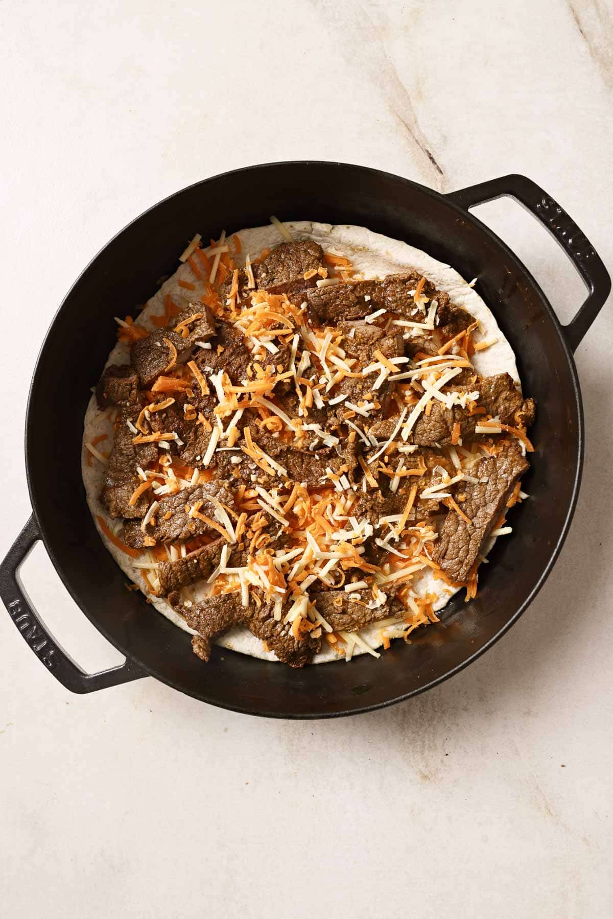 A cast iron skillet with a tortilla, cheese, and sliced carne asada.
