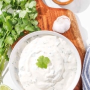 A bowl of cilantro lime crema on a wooden board, flanked by fresh cilantro, lime, garlic, and a pinch bowl of salt.