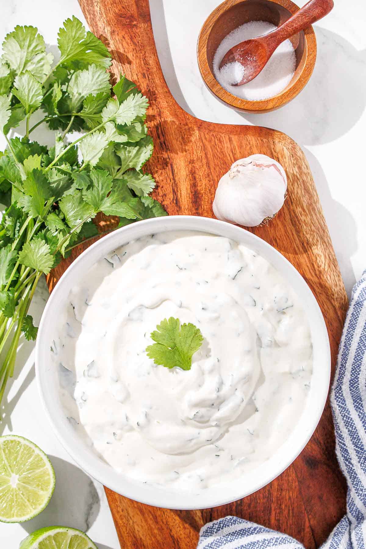 A bowl of cilantro lime crema on a wooden board, flanked by fresh cilantro, lime, garlic, and a pinch bowl of salt.