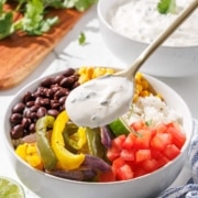 A colorful burrito bowl with a dollop of cilantro lime crema being added from a spoon.