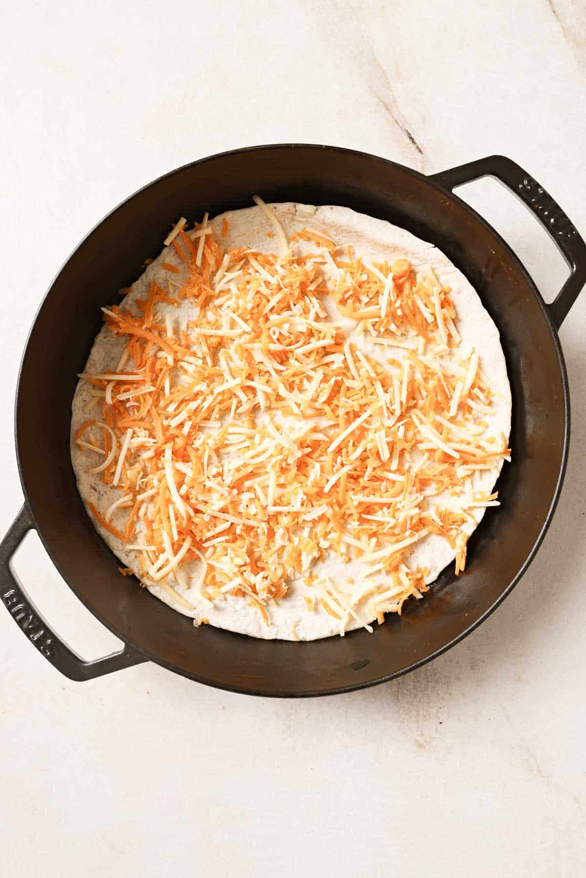 A cast iron skillet with a tortilla with shredded cheese.