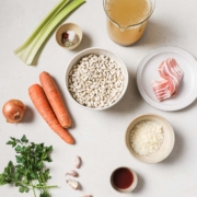 Ingredients for a recipe arranged on a light surface, including celery, beans, bacon, onion, carrots, parsley, garlic, rice, and broth.