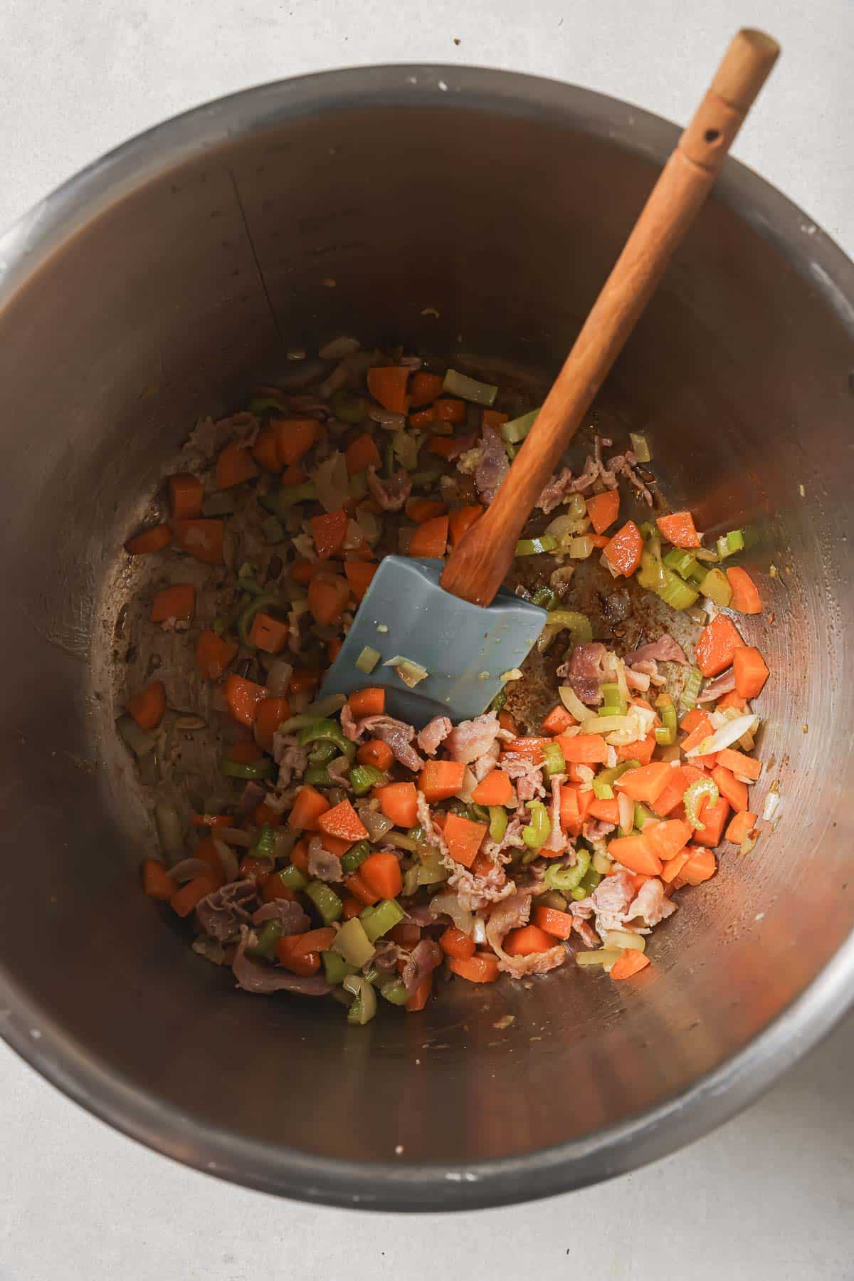 Chopped carrots, celery, and onion in a metal Instant pot with a wooden spatula.
