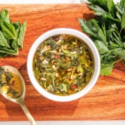 A bowl of fresh green chimichurri sauce on a wooden board, accompanied by herbs and a spoon.