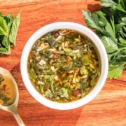 A bowl of mint chimichurri garnished with fresh parsley and mint leaves on a wooden board, accompanied by a spoon.