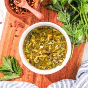 A bowl of mint chimichurri on a wooden board, garnished with fresh herbs and spices on the side.