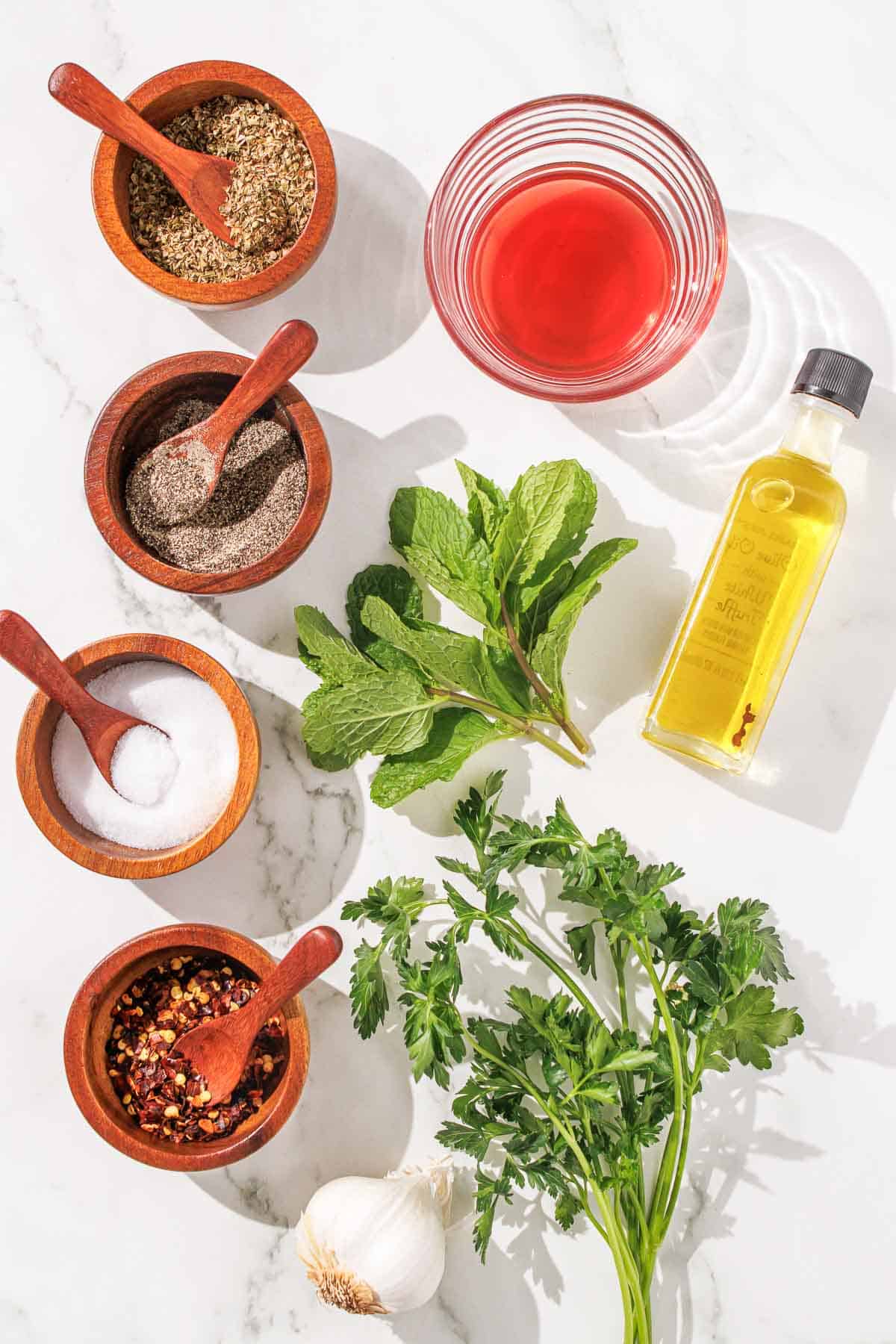 Overhead view of various cooking ingredients including herbs, spices, olive oil, and vinegar arranged neatly on a marble surface.