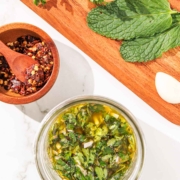 Overhead view of a homemade mint chimichurri in a glass jar, with spices in a wooden bowl and fresh herbs on a cutting board nearby.