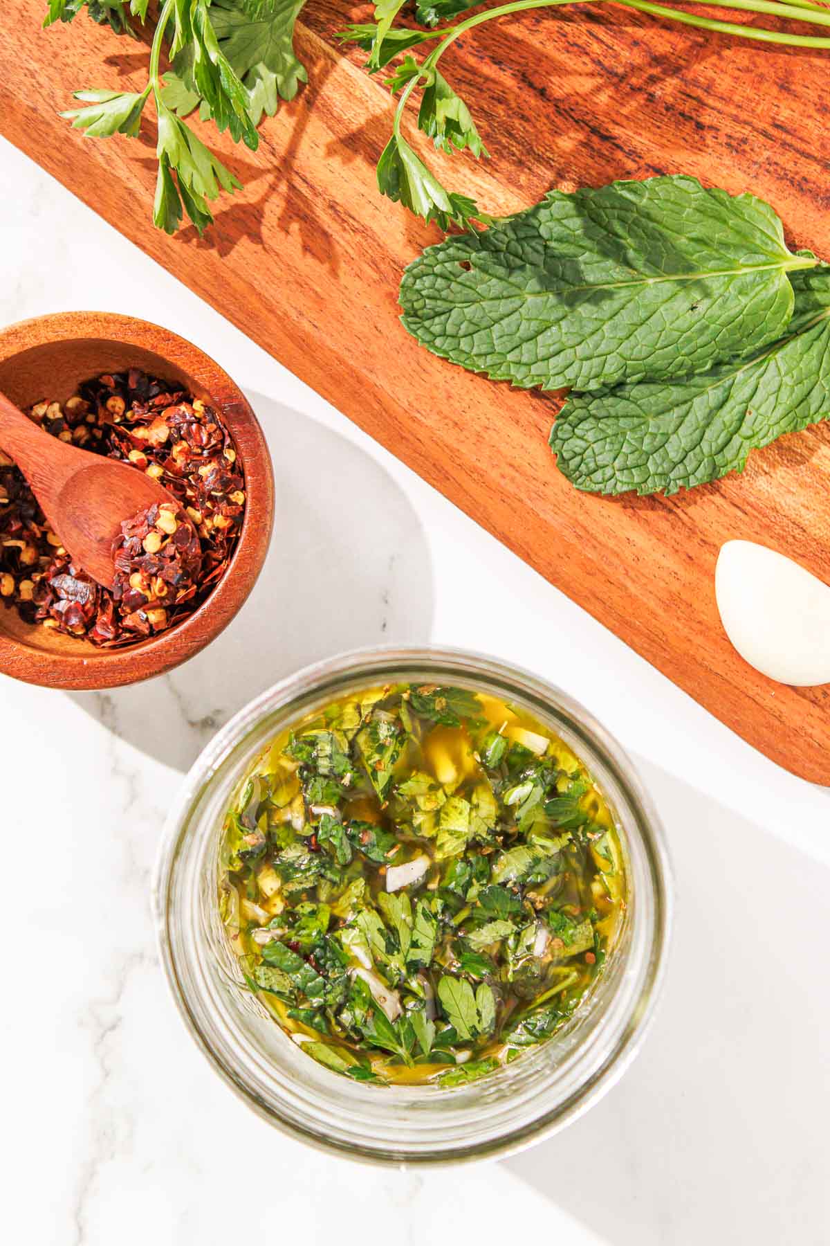 Overhead view of a homemade mint chimichurri in a glass jar, with spices in a wooden bowl and fresh herbs on a cutting board nearby.
