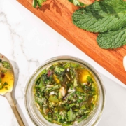 A glass jar filled with homemade chimichurri sauce on a marble counter, surrounded by fresh parsley and mint.