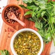 A bowl of mint chimichurri sauce on a wooden board with fresh herbs and a bowl of chili flakes beside it.