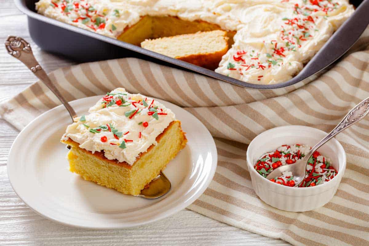 A slice of frosted yellow cake with red and green sprinkles on a plate. Nearby, a baking tray with more cake and a small dish of sprinkles on a striped cloth.