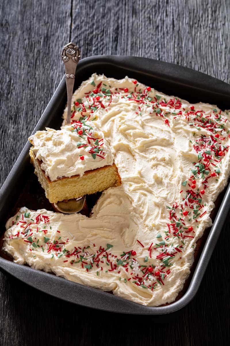 A rectangular frosted cake with holiday sprinkles in a baking pan, missing a corner piece on a serving spatula.