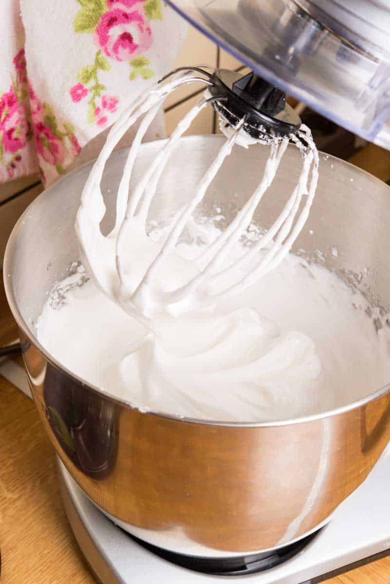 A stand mixer with a whisk attachment is shown whipping white cream in a stainless steel bowl. A floral patterned cloth is in the background.