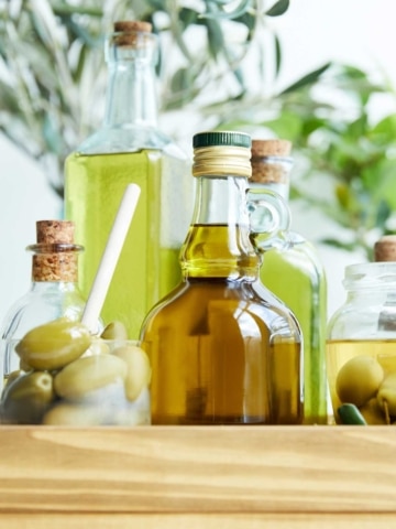 Several glass bottles filled with olive oil are arranged together, with jars of green olives and leafy olive branches in the background.